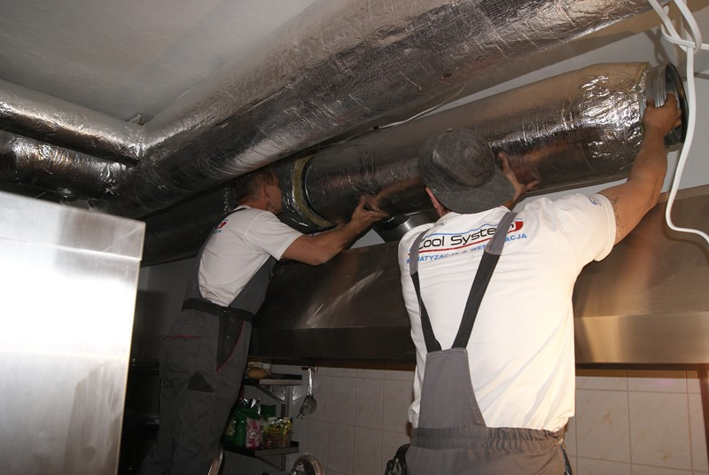 Dwóch monterów w kombinezonach firmowych instaluje izolowane kanały wentylacyjne pod sufitem w pomieszczeniu komercyjnym, widoczna stalowa obudowa okapu i rury wentylacyjne.