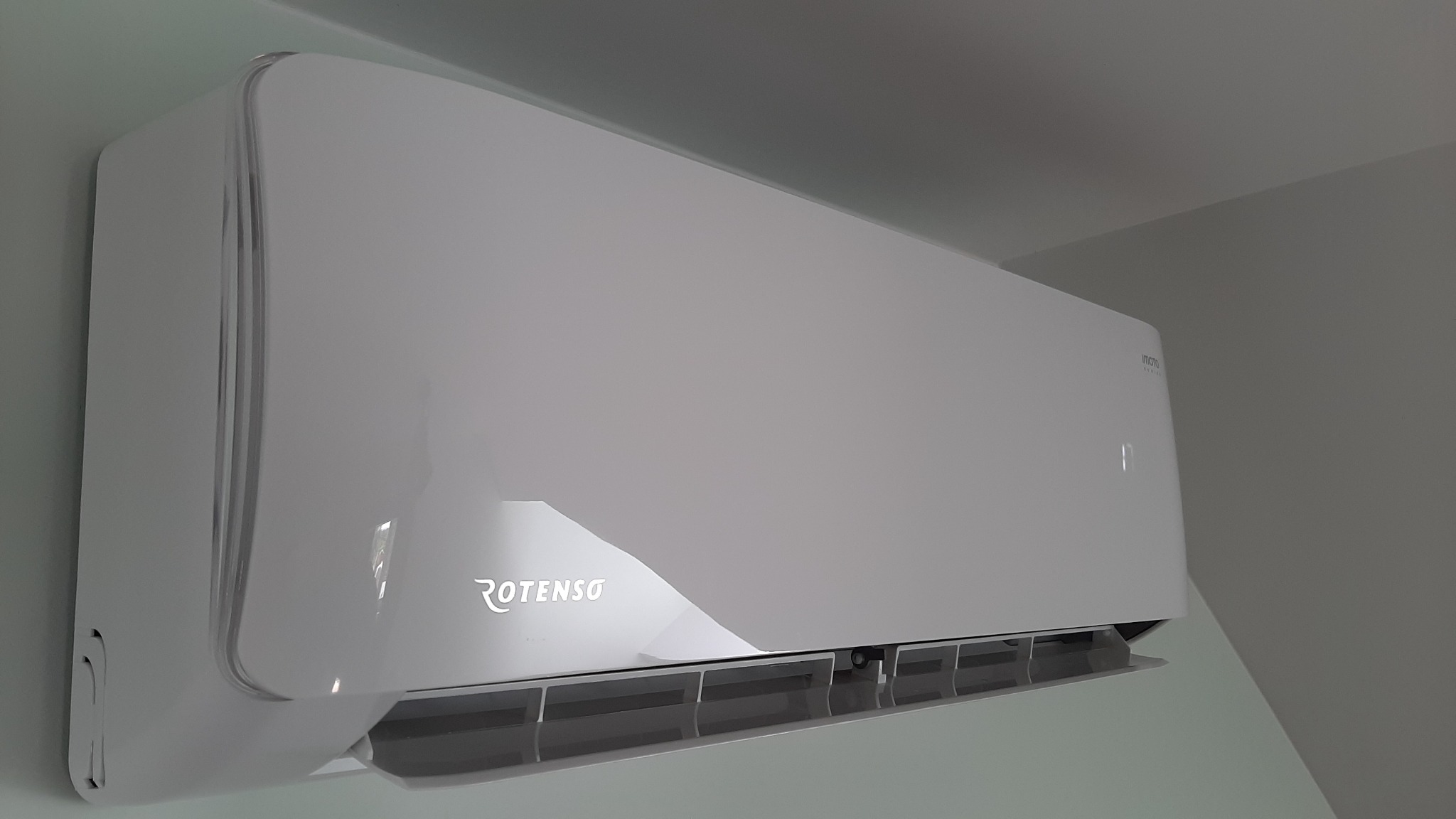 Biała jednostka klimatyzacji Rotenso zamontowana na ścianie, widoczna z boku, z otwartą klapką wylotu powietrza.