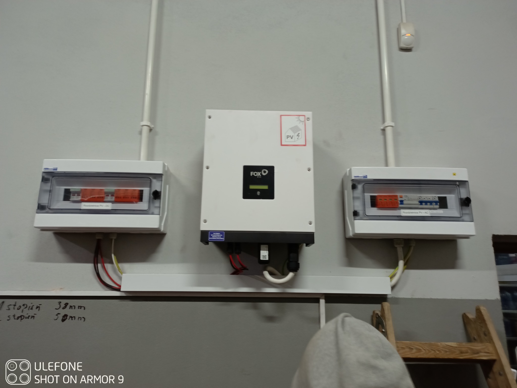 Zamontowany biały inwerter FOX z dwoma białymi rozdzielnicami po bokach na szarej ścianie, widoczne przewody i rurki instalacyjne, na dole fragment drabiny i czyjeś ramię.