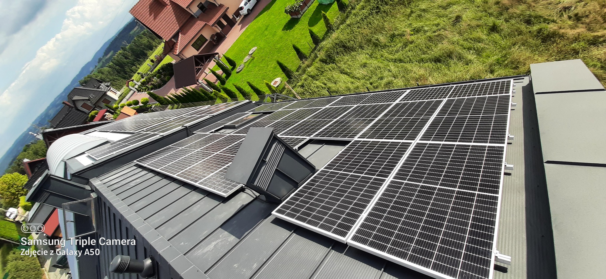 Instalacja paneli fotowoltaicznych na metalowym dachu domu jednorodzinnego, widok z góry, w tle zielony trawnik i budynki.