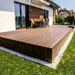 Nowoczesny taras z desek w odcieniach brązu, otoczony białym żwirem i zielonym trawnikiem, z widocznym zestawem mebli ogrodowych na tle ściany budynku z drewnianą okładziną.