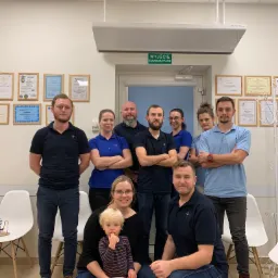 Zespół medyczny pozuje w gabinecie z dyplomami na ścianie, w tym kobieta z dzieckiem na kolanach, wszyscy ubrani w uniformy z logo.