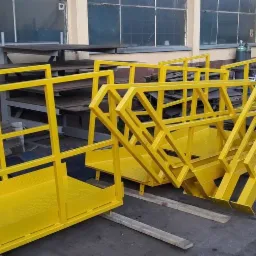 Żółte, metalowe konstrukcje schodowe z barierkami, prawdopodobnie do zastosowań przemysłowych, ustawione w warsztacie na drewnianych belkach, w tle okna i tabliczka informacyjna.