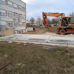 Michał Makowski - Pomarańczowa koparka kołowa obok betonowych płyt na trawniku, budynek w tle. Widoczne narzędzia i wykopany teren. Prace ziemne w toku.