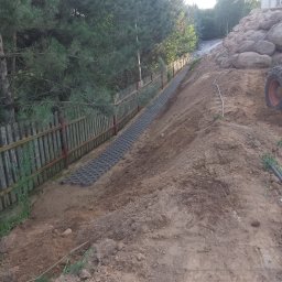 Michał Makowski - Ułożona, szara ażurowa nawierzchnia na zboczu, wzdłuż drewnianego płotu z czerwonymi słupkami. W tle drzewa i kamienny mur oporowy.