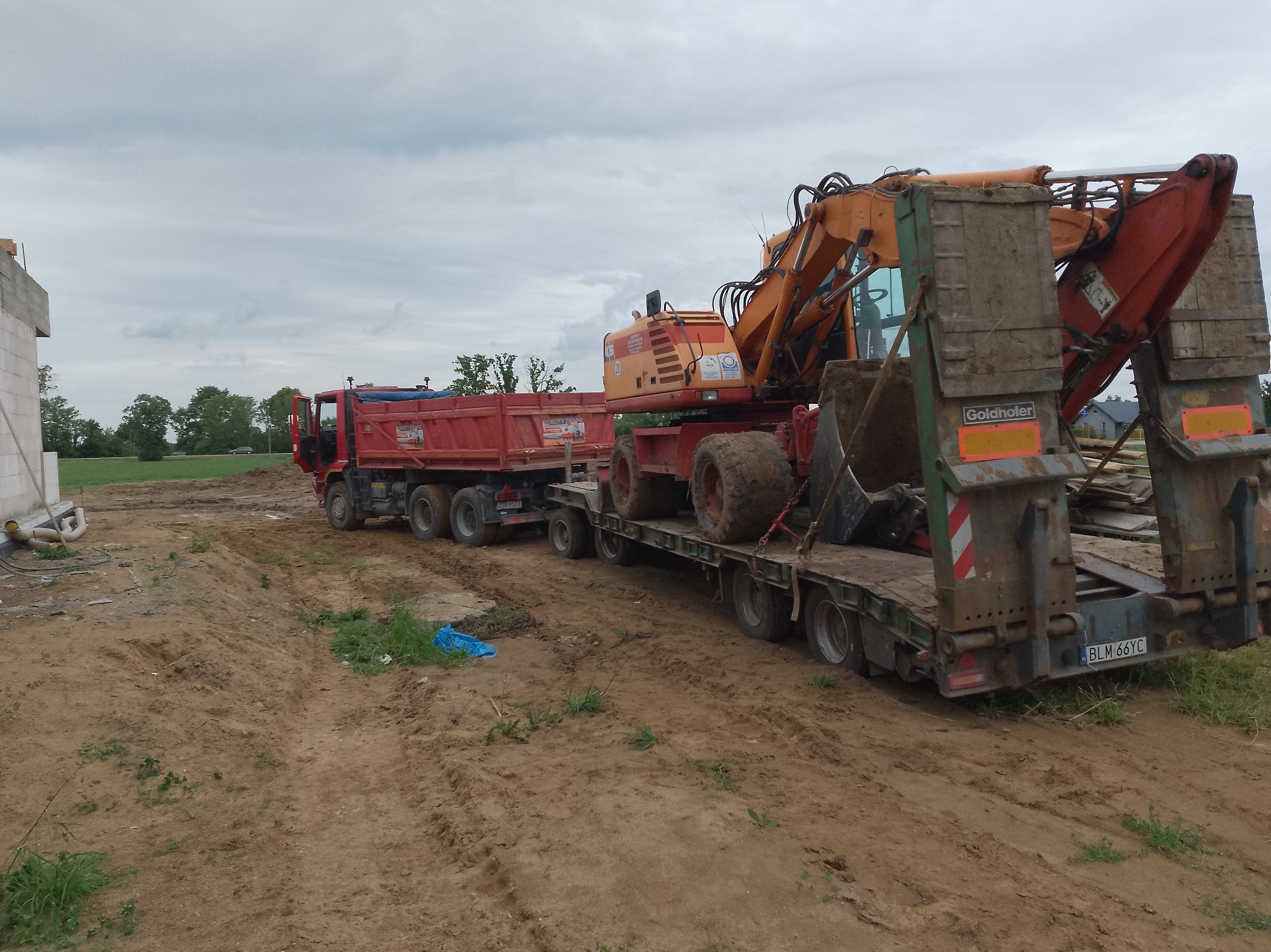 Ciężarówka z naczepą transportuje pomarańczową koparkę na placu budowy. W tle widoczny budynek w trakcie budowy i zielone drzewa pod pochmurnym niebem.