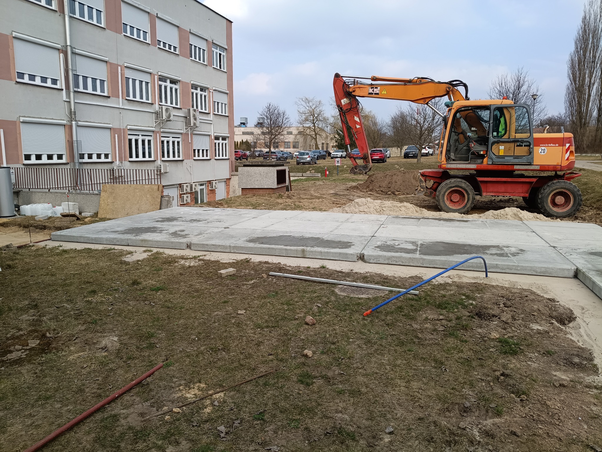 Pomarańczowa koparka kołowa obok betonowych płyt na trawniku, budynek w tle. Widoczne narzędzia i wykopany teren. Prace ziemne w toku.