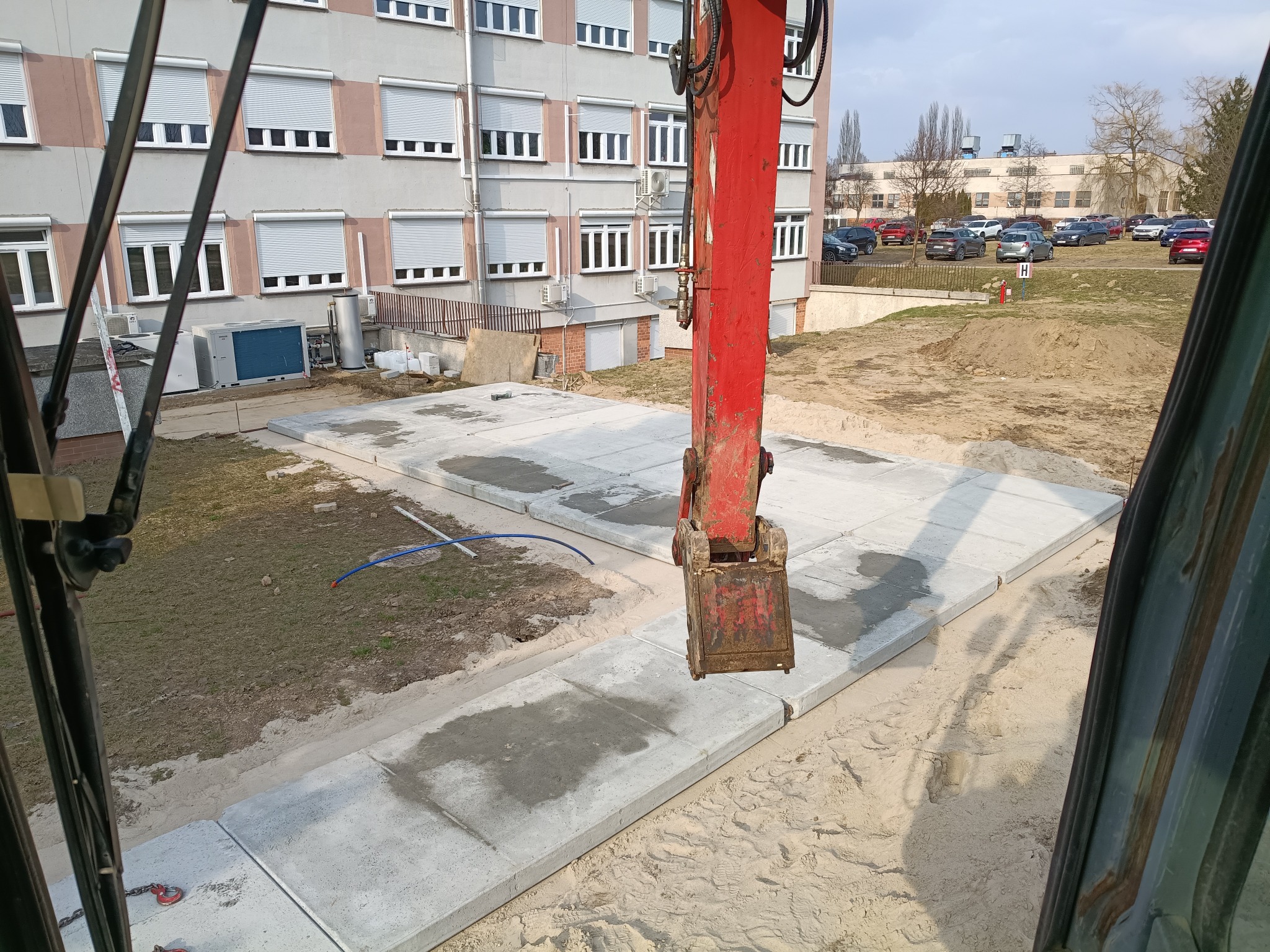 Częściowo ułożone płyty chodnikowe na piasku, widok z kabiny koparki. W tle budynek z licznymi oknami i parking. Elementy koparki w kadrze.