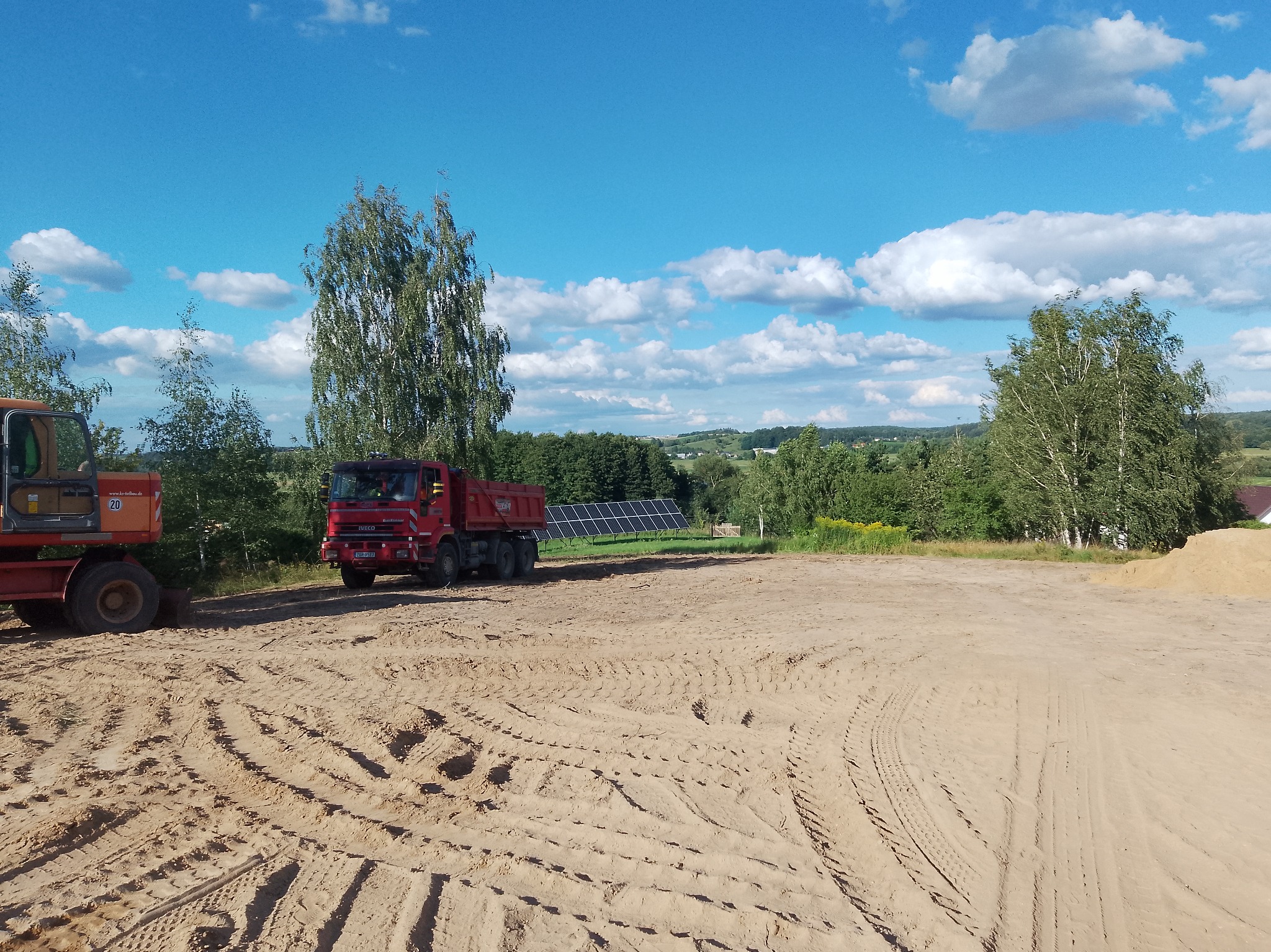 Ciężarówka i koparka na placu budowy w Łomży, przygotowanym pod prace ziemne. W tle widoczne panele słoneczne i zieleń.