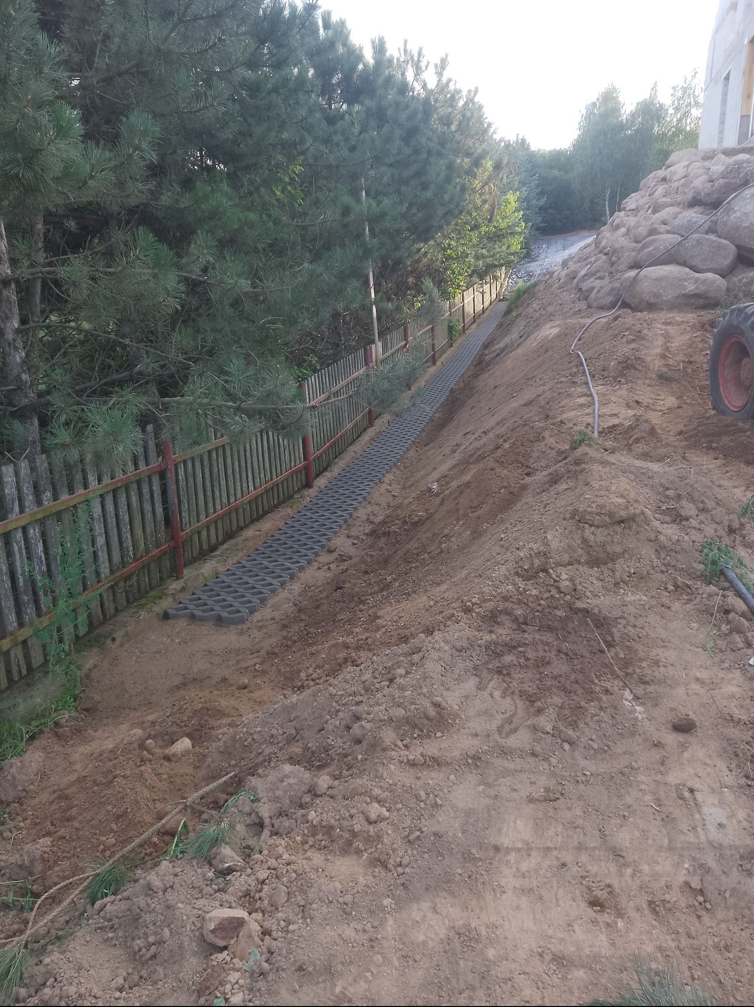 Ułożona, szara ażurowa nawierzchnia na zboczu, wzdłuż drewnianego płotu z czerwonymi słupkami. W tle drzewa i kamienny mur oporowy.