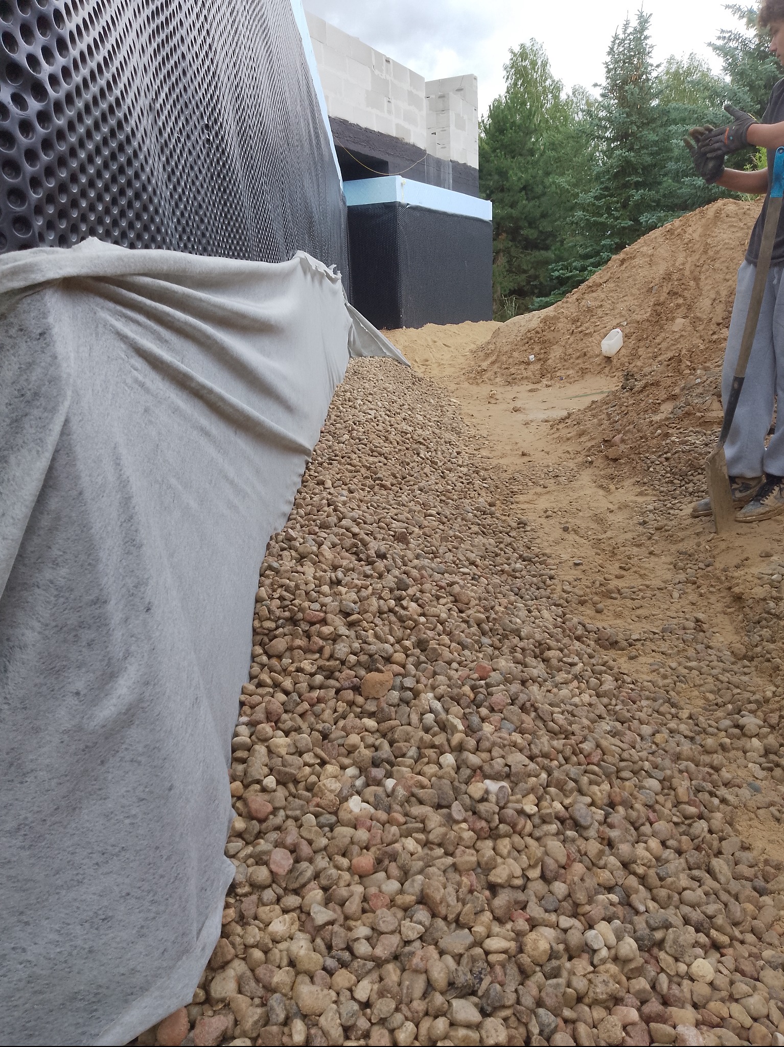 Ocieplenie fundamentów budynku z użyciem folii kubełkowej i geowłókniny, obsypane drobnym żwirem. Widoczny fragment pracownika z łopatą.