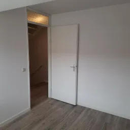 Wnętrze pokoju po odświeżeniu: białe ściany, drewnopodobna podłoga, otwarte drzwi z widokiem na schody z białymi poręczami, nadproże z dekoracyjnym szkłem.