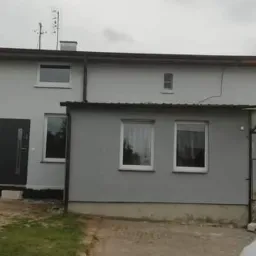 Świeżo pomalowana szara elewacja szeregowego domu z ciemną stolarką okienną i drzwiową, widoczny fragment trawnika i materiały budowlane.