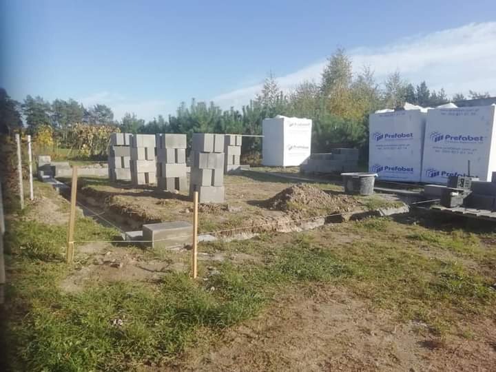 Fundamenty domu w trakcie budowy: wykopane rowy, ustawione bloczki betonowe, widoczne palety z materiałami budowlanymi Prefabet, teren trawiasty.