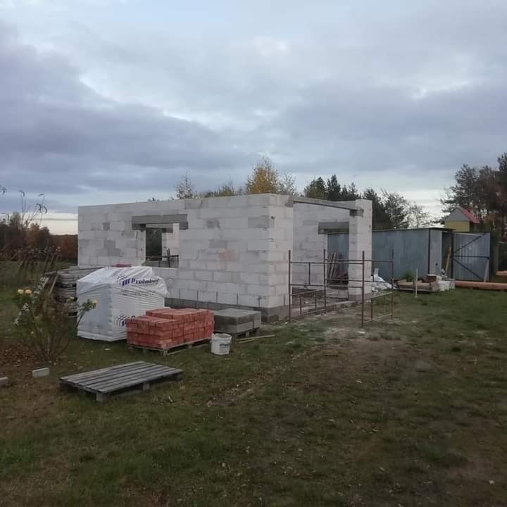 Budowa domu jednorodzinnego w stanie surowym otwartym, ściany z białego betonu komórkowego, widoczne palety z bloczkami, cegłą czerwoną i rusztowanie, na tle ponurego nieba i drzew.