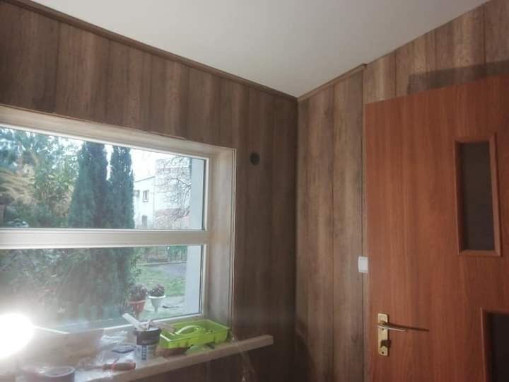 Pokój wykończony panelami ściennymi imitującymi drewno, widoczne okno z parapetem, na którym leżą narzędzia i akcesoria malarskie, oraz brązowe drzwi z przeszkleniem.