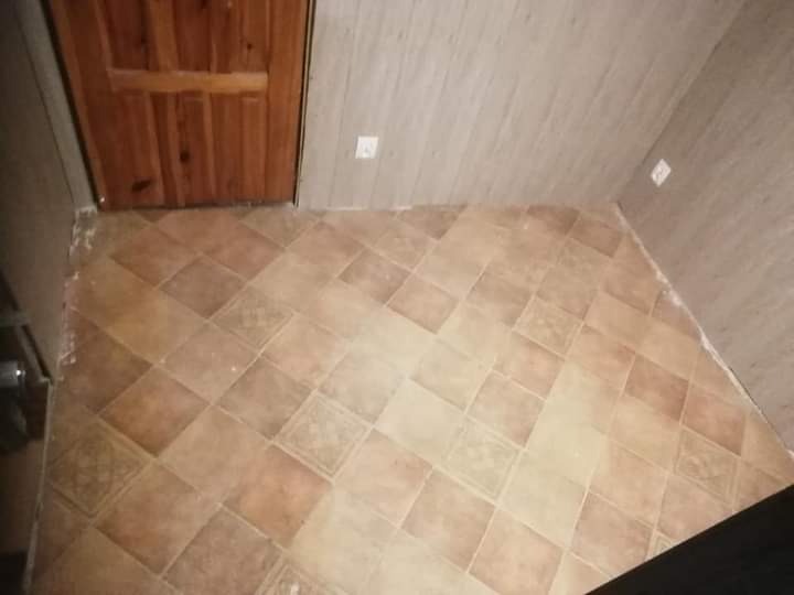 Kafelkowana podłoga w odcieniach beżu i brązu ułożona w romby, drewniane drzwi w tle, ściany wykończone panelami w kolorze beżowym.