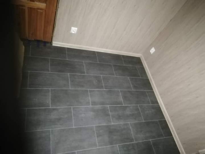 Wykończona podłoga z szarych, prostokątnych płytek w pomieszczeniu z panelami ściennymi w odcieniach beżu i białym wykończeniem przy podłodze i suficie, widoczne gniazdko elektryczne.