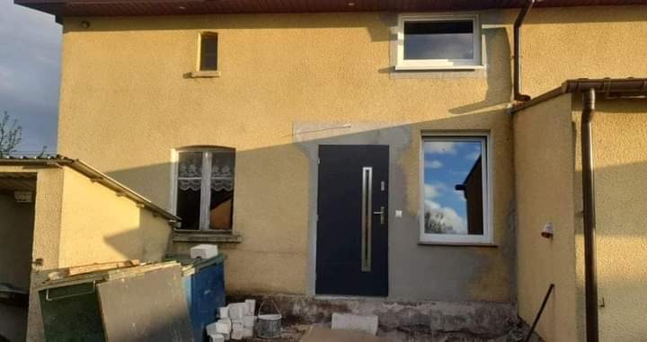 Elewacja domu z nowymi oknami PCV i drzwiami zewnętrznymi w kolorze grafitowym, widoczne ślady po pracach wykończeniowych, w tle niebieskie niebo odbijające się w szybach.