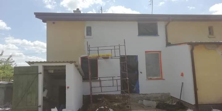 Dwukondygnacyjny dom w trakcie malowania elewacji, widoczne rusztowania, fragmenty pomalowane na biało, a inne jeszcze w kolorze piaskowym, z otwartymi drzwiami do garażu.