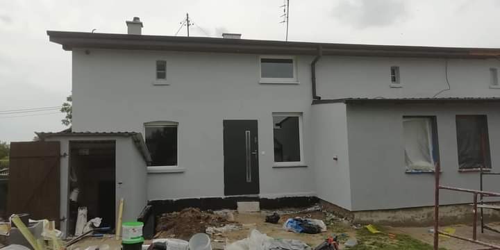 Szary bliźniak po malowaniu elewacji, widoczne przygotowania do dalszych prac wykończeniowych, folia na oknach, niezagospodarowany teren przed budynkiem.