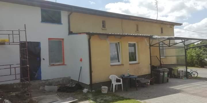 Elewacja domu w trakcie odnawiania: jedna część pomalowana na biało, druga w kolorze żółtym, widoczne rusztowanie, okna zabezpieczone folią, elementy wyposażenia posesji. Prace w toku.