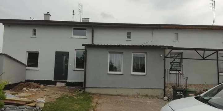 Świeżo pomalowana szara elewacja szeregowego domu z ciemną stolarką okienną i drzwiową, widoczny fragment trawnika i materiały budowlane.