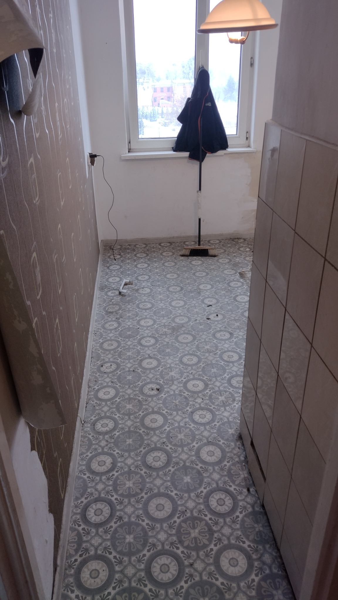 Wnętrze w trakcie remontu: położona wzorzysta szaro-biała podłoga, ściana częściowo z tapetą, okno z kurtką i mopem, widoczne ślady prac remontowych.