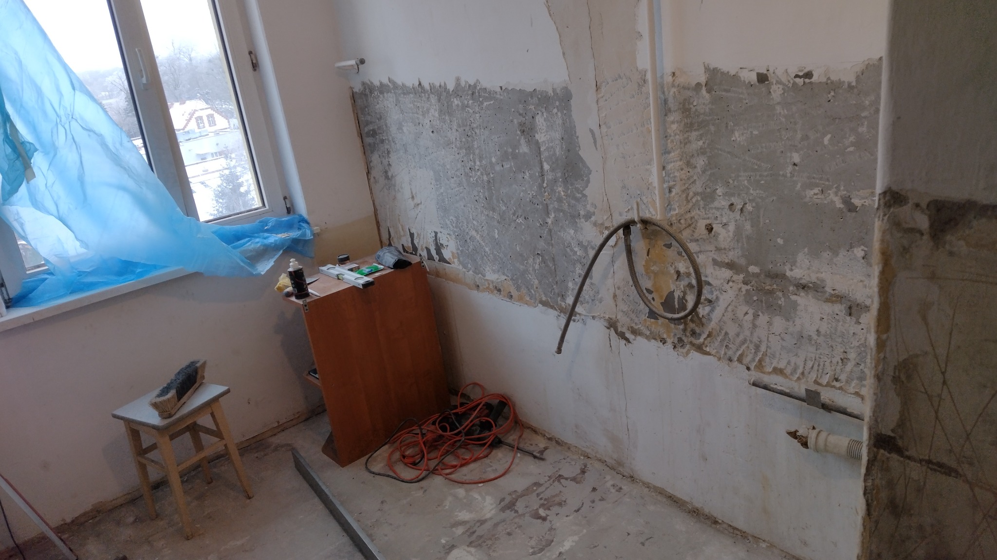 Pokój w trakcie remontu: ściany częściowo skuite, widoczne instalacje, okno zabezpieczone folią, narzędzia i szafka w pomieszczeniu.