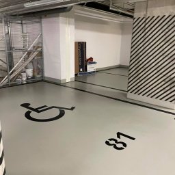 Iw-Bud - Oznakowane miejsce parkingowe dla osób niepełnosprawnych w garażu podziemnym, numer 81, widoczne rury pod sufitem i regał z farbami w tle.