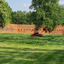 Iw-Bud - Mężczyzna na czerwonej kosiarce traktorowej kosi wysoką trawę na tle ceglanego muru z otworami strzelniczymi i zielonych drzew.