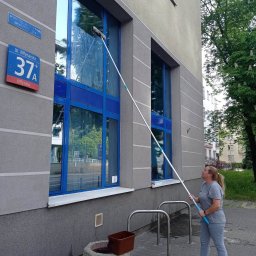 Iw-Bud - Kobieta myje okna na wysokości przy użyciu długiego kija z mopem, widoczny budynek z niebieskimi ramami okiennymi i tabliczka z adresem 'Ul. Młynarska 37A'.