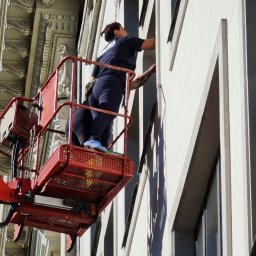 Iw-Bud - Dwie osoby w podnośniku koszowym czyszczą okna na wysokości w budynku z dekoracyjną fasadą.