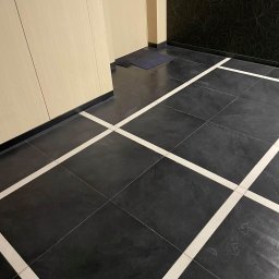Iw-Bud - Ciemne płytki podłogowe z białymi fugami krzyżującymi się, widoczne fragmenty ścian z kremowym i czarnym wykończeniem oraz drzwi.