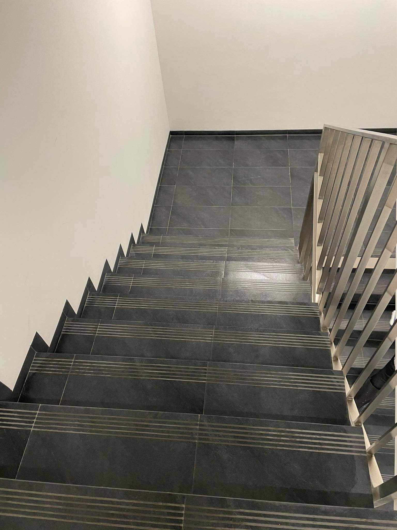 Klatka schodowa wyłożona ciemnymi płytkami z poziomymi paskami antypoślizgowymi, widok z góry, metalowa balustrada po prawej stronie.