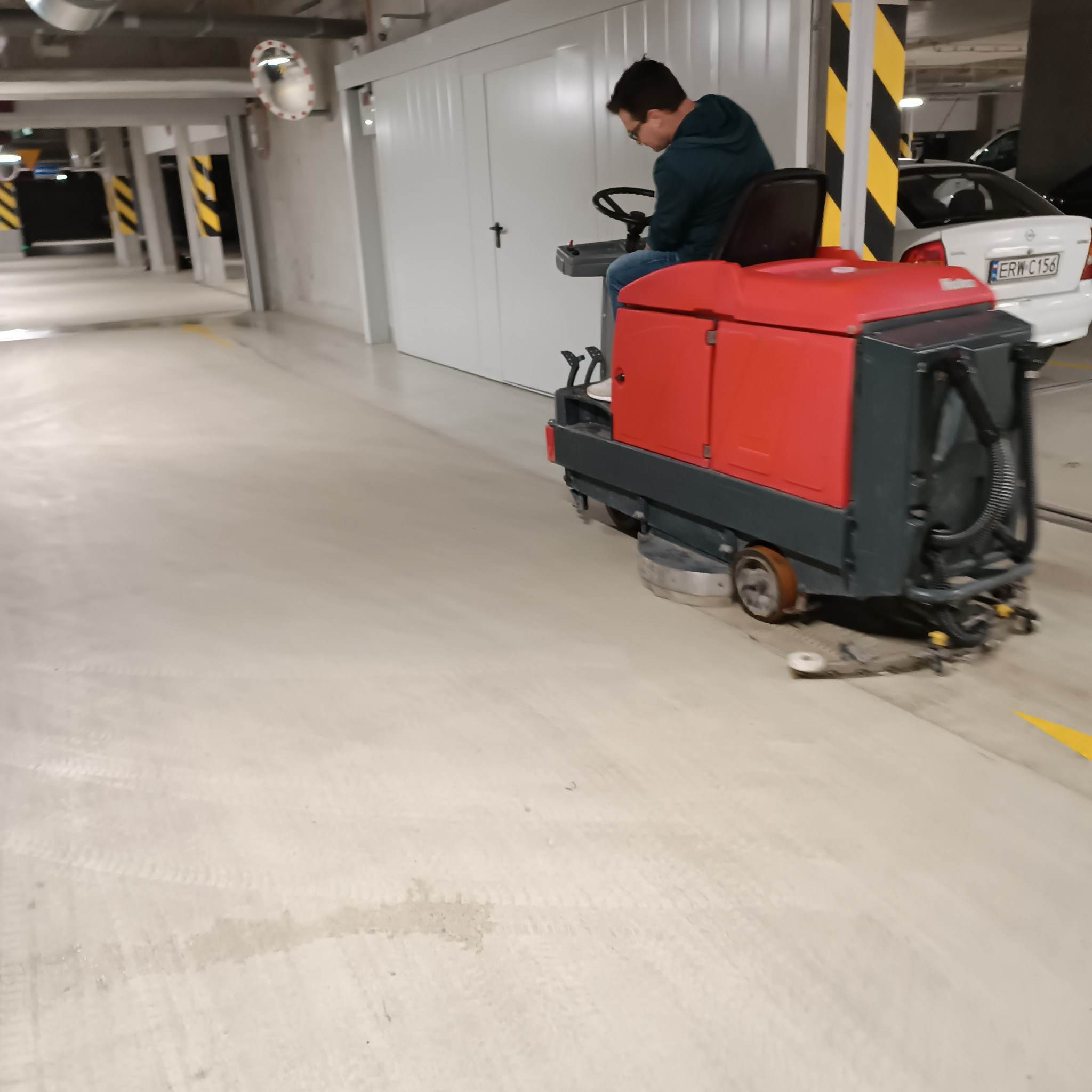 Operator na maszynie czyszczącej podłogę w garażu podziemnym, widoczne ślady po czyszczeniu.
