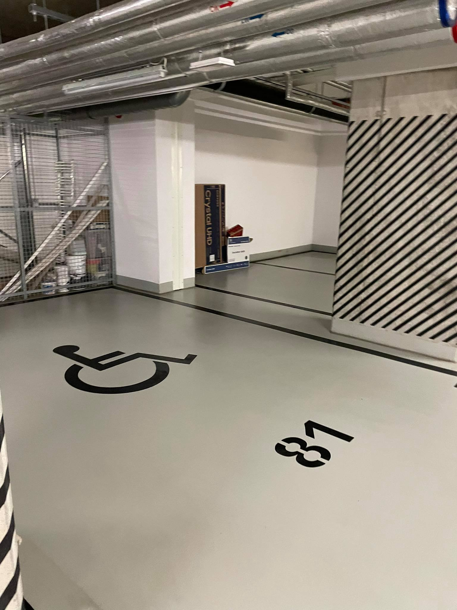 Oznakowane miejsce parkingowe dla osób niepełnosprawnych w garażu podziemnym, numer 81, widoczne rury pod sufitem i regał z farbami w tle.