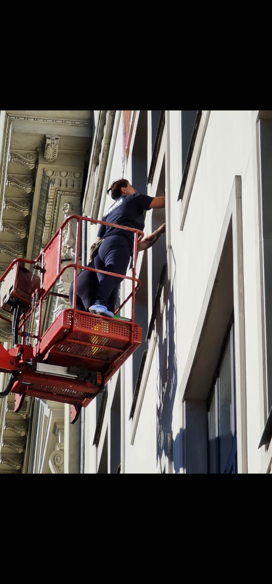 Dwie osoby w podnośniku koszowym czyszczą okna na wysokości w budynku z dekoracyjną fasadą.