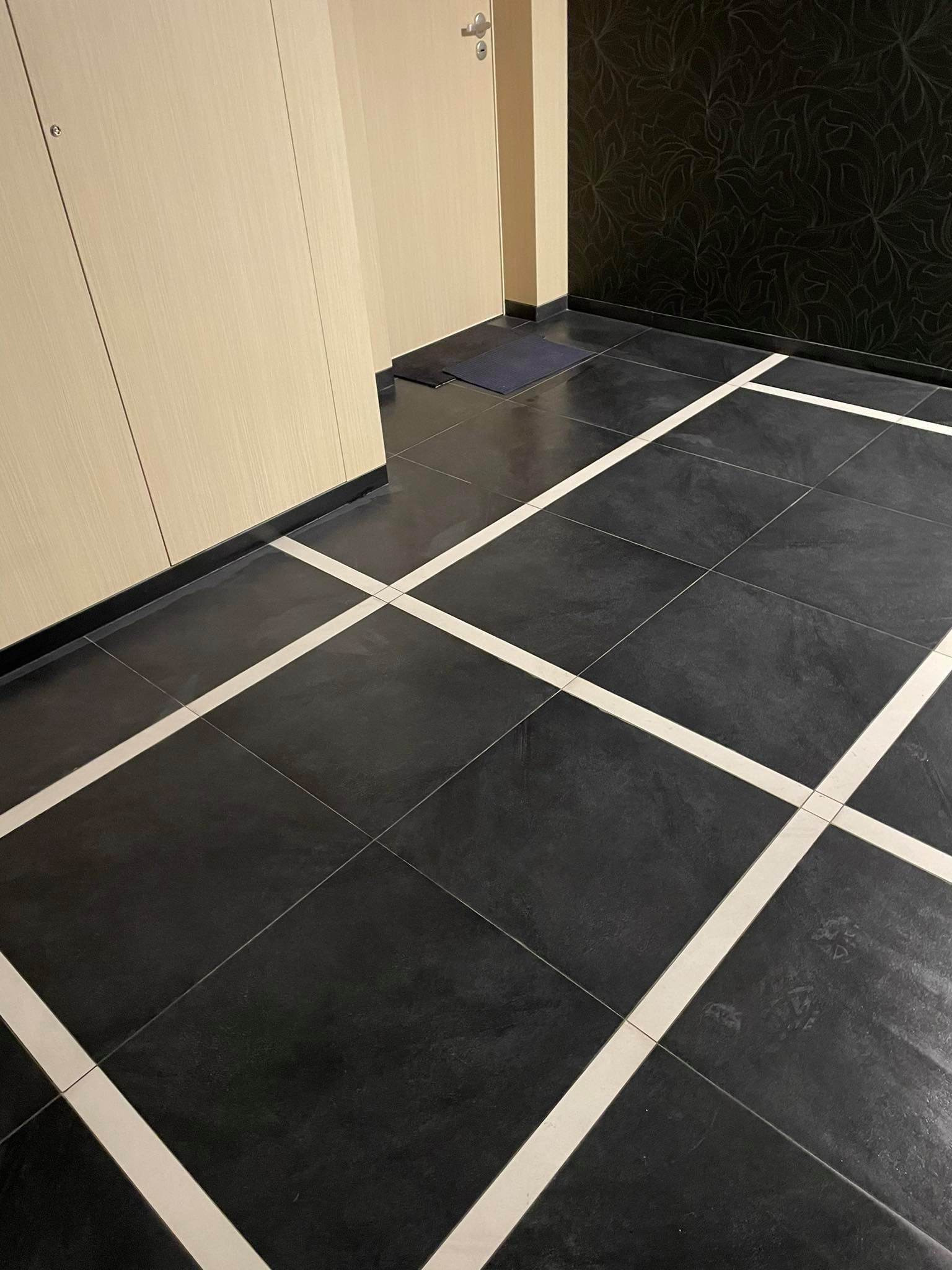 Ciemne płytki podłogowe z białymi fugami krzyżującymi się, widoczne fragmenty ścian z kremowym i czarnym wykończeniem oraz drzwi.