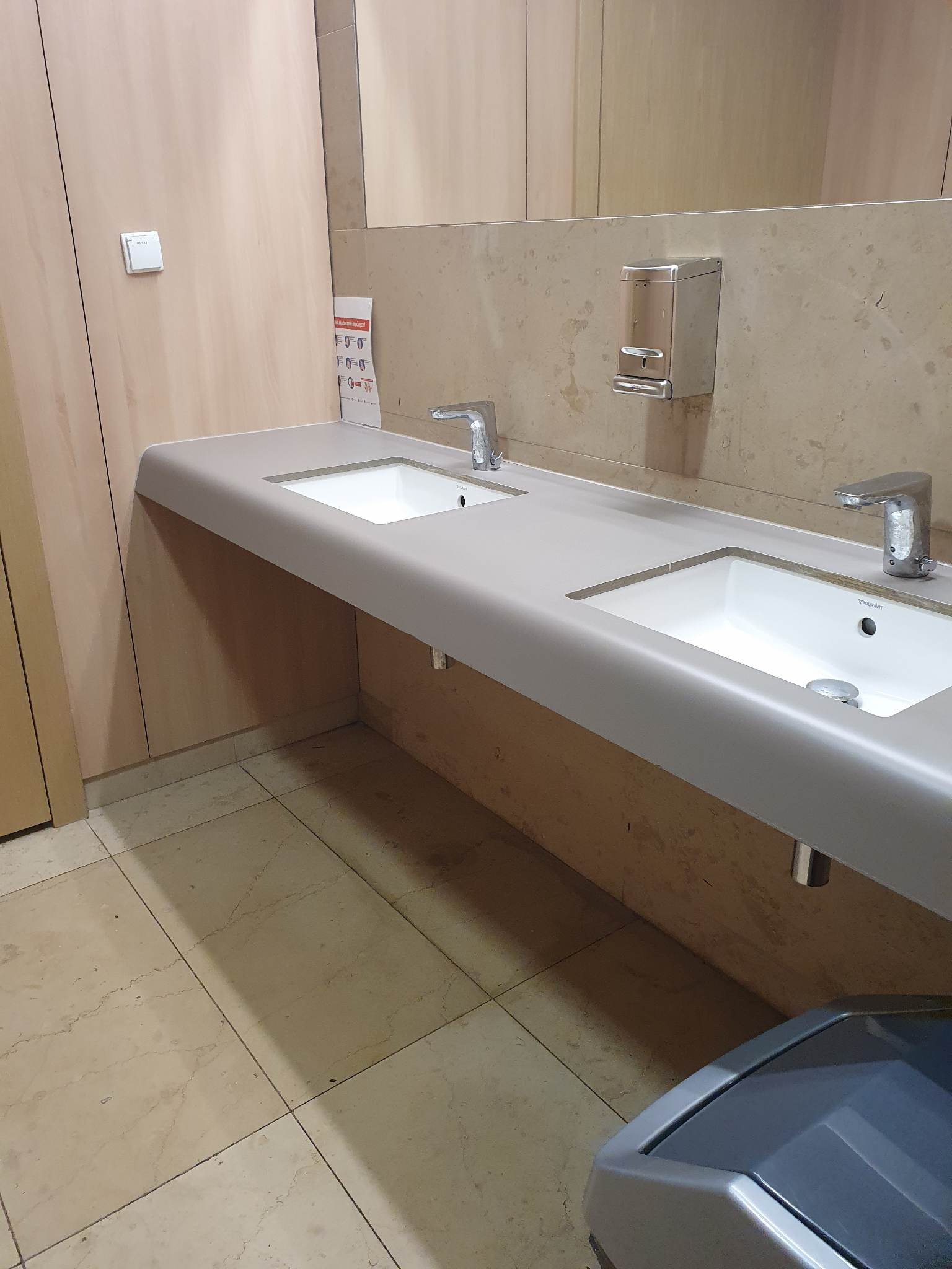 Dwie białe umywalki z chromowanymi kranami na szarym blacie, zamontowane w publicznej toalecie z beżowymi płytkami podłogowymi i ścianami w odcieniach beżu i brązu, z widocznym dozownikiem mydła...