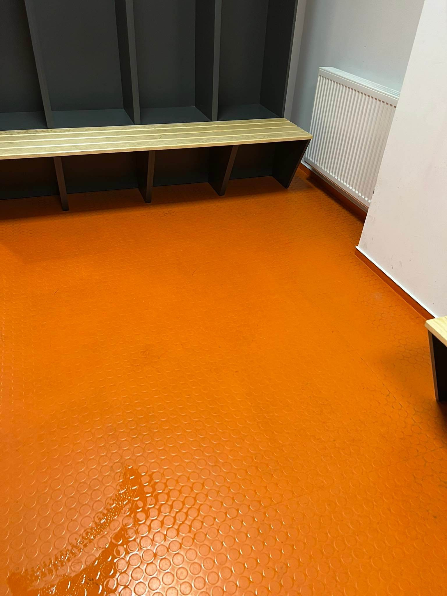 Pomarańczowa podłoga z wypustkami w szatni, widoczne szafki z ławką i grzejnik, ślady wilgoci na podłodze.