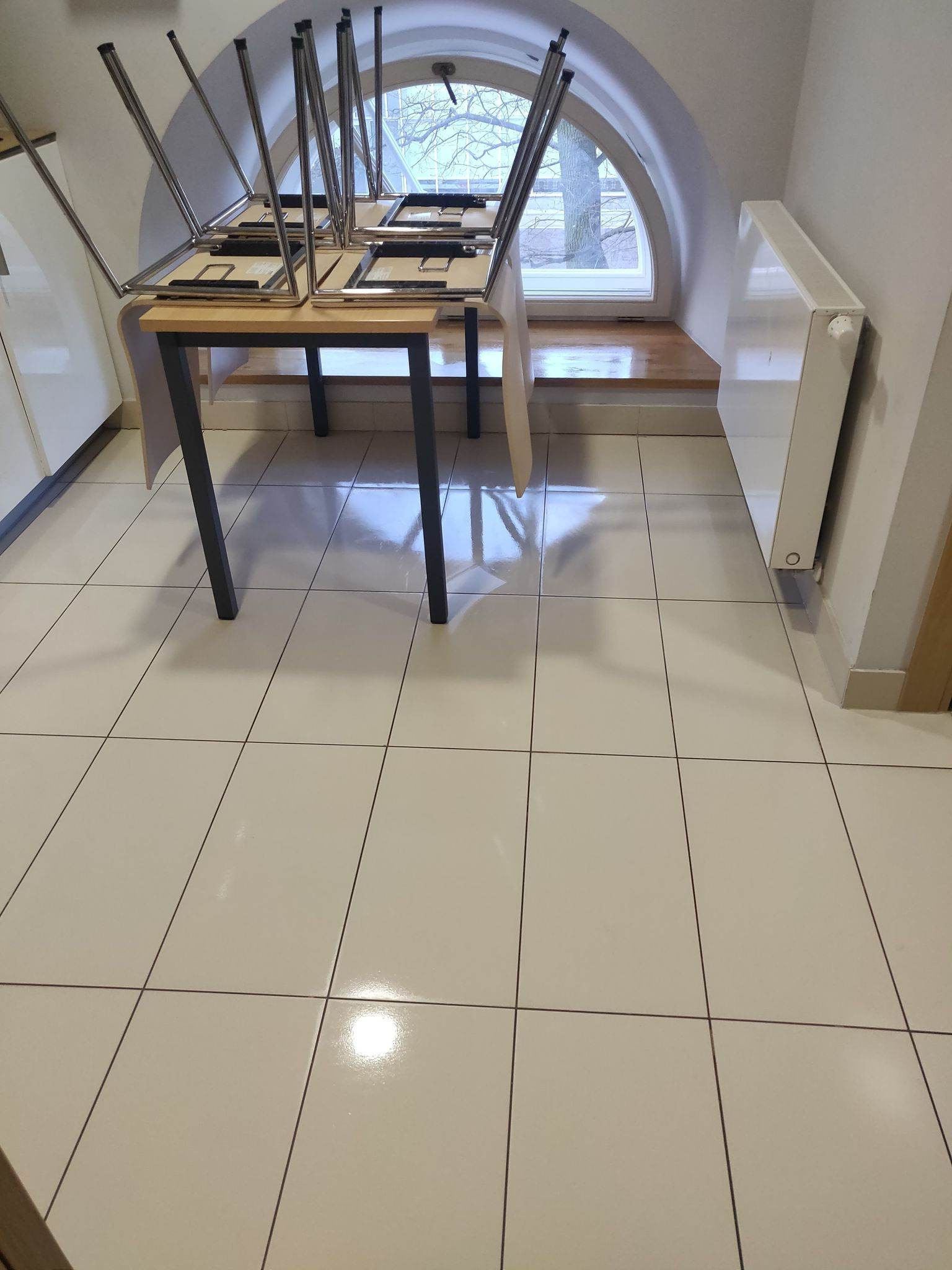 Ułożone krzesła na stole w pustym pomieszczeniu z błyszczącą, jasną podłogą z płytek i oknem w kształcie łuku.