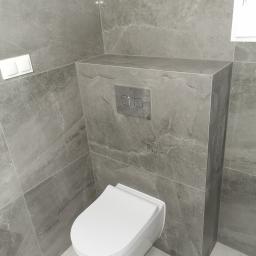 Artur Rodak Remonty i wykonczenia - Nowoczesna łazienka z zabudowaną toaletą w szarych płytkach imitujących marmur, widoczne chromowane przyciski spłukiwania i biała miska WC.