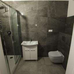 Artur Rodak Remonty i wykonczenia - Nowoczesna łazienka z szarymi płytkami imitującymi marmur, kabina prysznicowa ze szklanymi drzwiami przesuwnymi, podwieszana umywalka z szafką i wisząca toaleta.