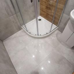 Artur Rodak Remonty i wykonczenia - Nowoczesna łazienka po remoncie z narożną kabiną prysznicową z chromowanymi profilami, drewnopodobnymi płytkami wewnątrz kabiny, szarą płytką podłogową z połyskiem i butami w dolnym kadrze.