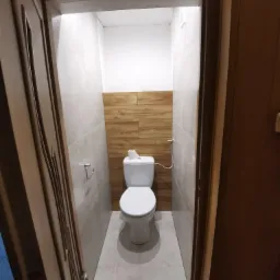 Wnętrze małej toalety po remoncie, widok zza uchylonych, zdobionych drzwi. Ściany wyłożone szarymi płytkami, fragment ściany nad toaletą w okładzinie imitującej drewno, biała toaleta kompaktowa...