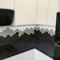 Fragment nowoczesnej kuchni z białymi szafkami, czarnym blatem i heksagonalnymi płytkami w odcieniach szarości na ścianie, widoczny czarny okap i piekarnik.