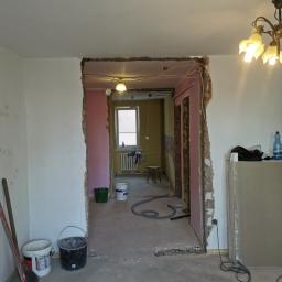 Artur Rodak Remonty i wykonczenia - Przygotowania do prac wykończeniowych: widoczne ślady po kuciu ścian, odkryte przewody elektryczne, wiadro z zaprawą i płyty gipsowo-kartonowe w pomieszczeniu przed remontem.