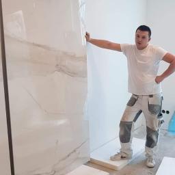 Artur Rodak Remonty i wykonczenia - Instalator opiera się o dużą, błyszczącą płytę imitującą marmur, ustawioną pionowo na podłodze wyłożonej styropianem w trakcie prac wykończeniowych w jasnym pomieszczeniu.