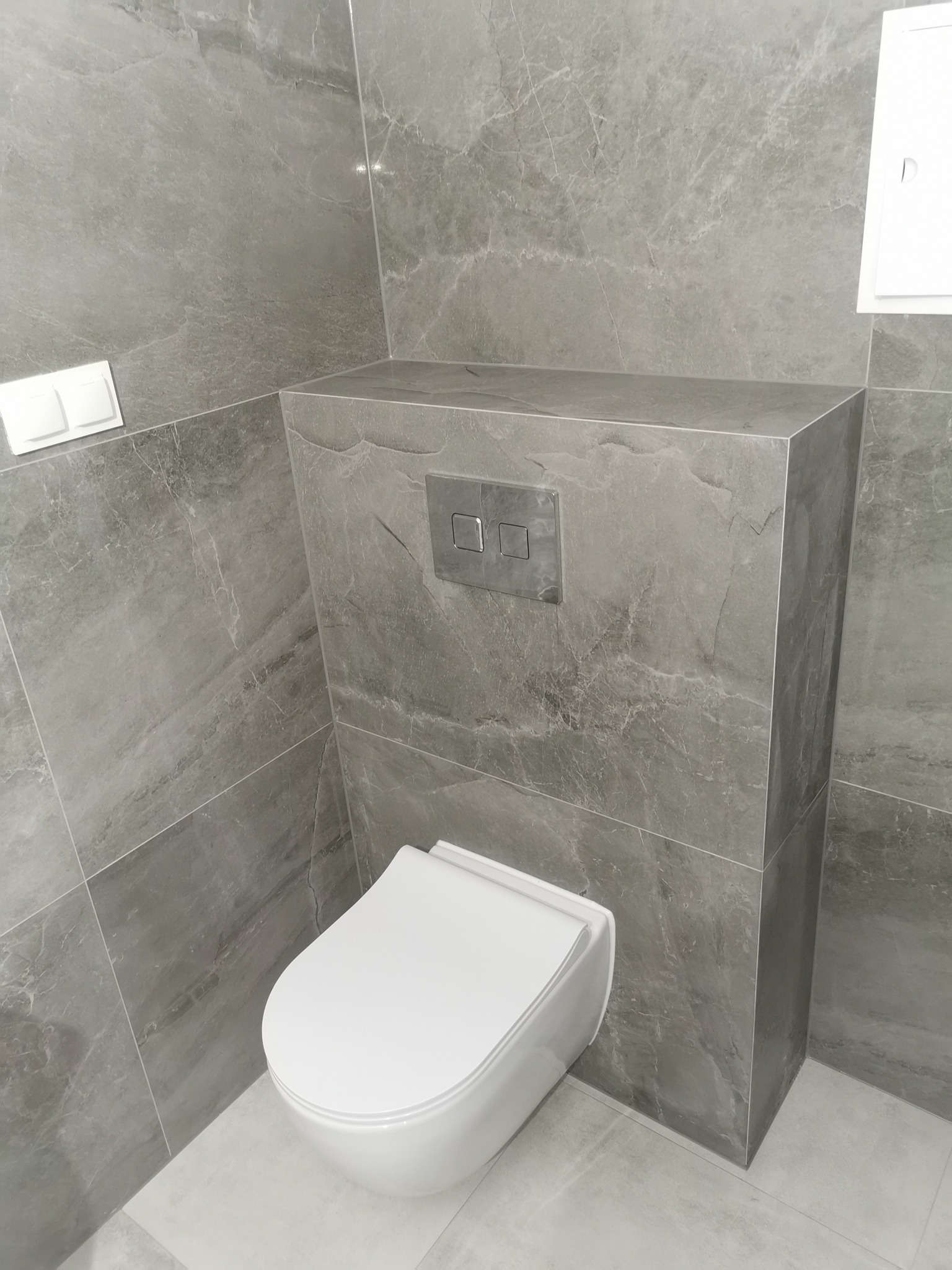 Nowoczesna łazienka z zabudowaną toaletą w szarych płytkach imitujących marmur, widoczne chromowane przyciski spłukiwania i biała miska WC.